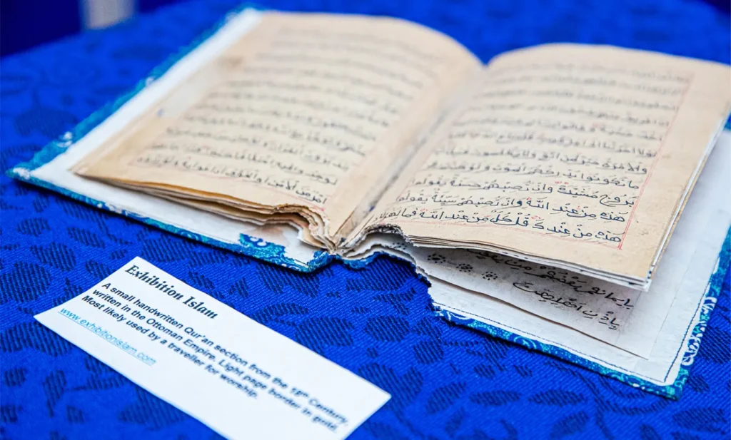 An open handwritten Qur'an on blue cloth with an Exhibition Islam label